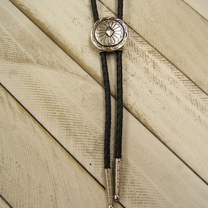 May include: A black bolo tie with a silver, circular, decorative tip featuring a geometric design. The bolo tie is on a white, wood background.
