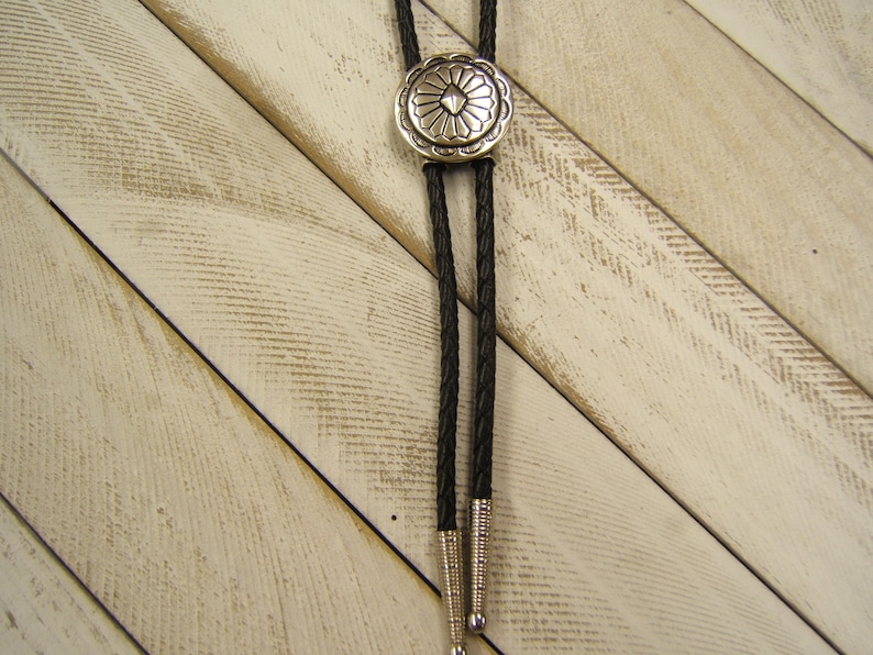 May include: A black bolo tie with a silver, circular, decorative tip featuring a geometric design. The bolo tie is on a white, wood background.
