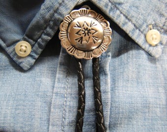 May include: A silver bolo tie with a decorative circular pendant featuring an engraved starburst design. The pendant is attached to a braided black leather cord, set against a light blue button-down shirt.