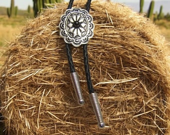 May include: A silver bolo tie with a decorative, ornate, circular design. The tie is on a bale of hay.