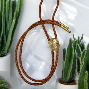 May include: A brown leather bolo tie with a gold-toned metal tip featuring a floral design. The bolo tie is displayed against a white background with green cacti.