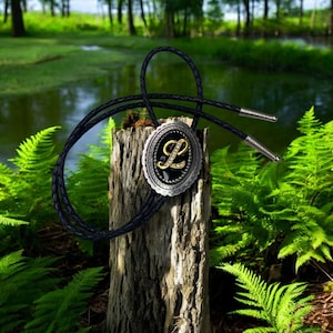 May include: A black bolo tie with a silver and gold bolo tie slide featuring the letter L with rhinestones. The bolo tie is on a tree stump in a natural setting.