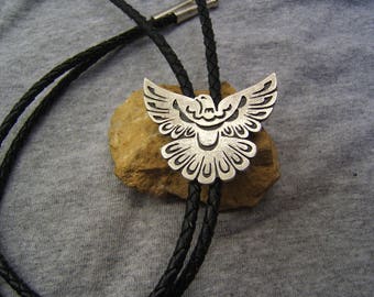 May include: A black bolo tie with a silver dove pendant. The dove is detailed with intricate designs and has a spread-wing pose.