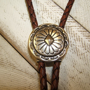 May include: A brown leather bolo tie with a silver, circular, decorative tip featuring a geometric design. The tip has a raised, diamond-shaped center surrounded by a radiating pattern.