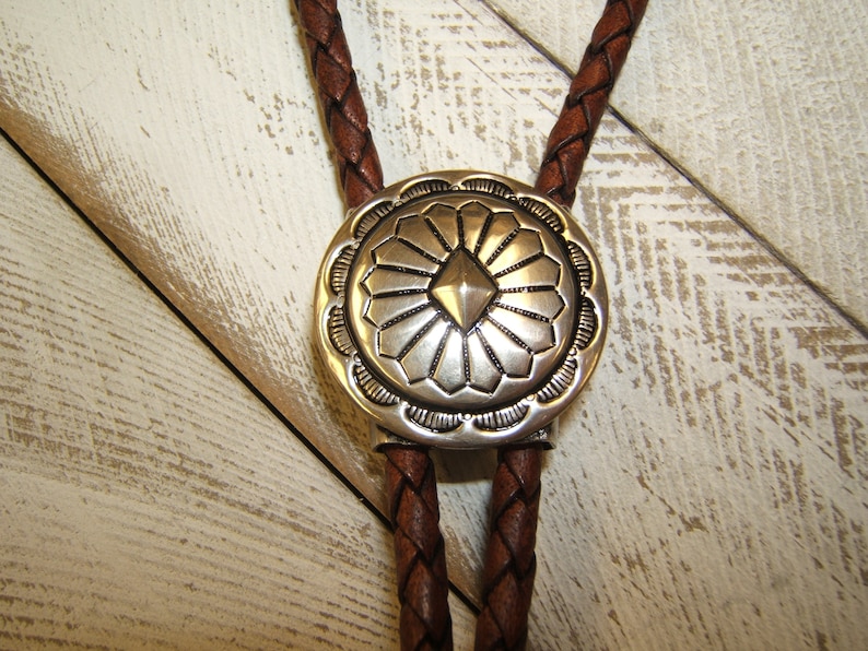 May include: A brown leather bolo tie with a silver, circular, decorative tip featuring a geometric design. The tip has a raised, diamond-shaped center surrounded by a radiating pattern.