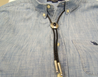 May include: A black bolo tie with a silver and black bolo tie slide. The slide has a circular design with a black stone in the center.