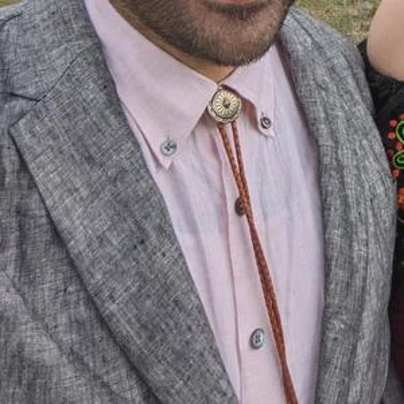 May include: A bolo tie with a circular, ornate, gold-colored metal pendant. The tie has a brown leather cord and is worn over a light pink button-down shirt and a gray blazer.
