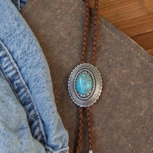 May include: A bolo tie featuring a central oval turquoise stone set in a decorative silver-toned metal frame. The tie consists of a braided brown leather cord finished with two silver-toned metal tips at the ends of the cord strands.