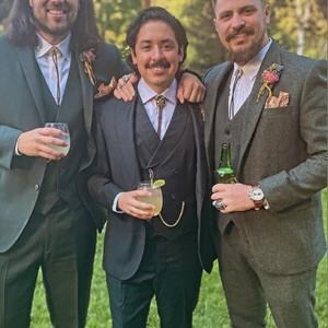 May include: Three men in suits pose outdoors. The suits vary in color, including dark green, charcoal, and a textured gray. Each man wears a vest, and two have bolo ties. One holds a glass of a beverage, and another holds a bottle.