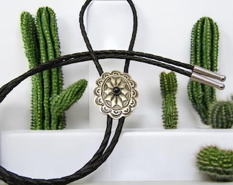 May include: A black braided bolo tie with a silver concho featuring a black stone in the center. The concho has a decorative design with a starburst pattern.