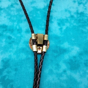 May include: A black braided bolo tie with a gold-toned metal clasp. The tie is on a turquoise background.