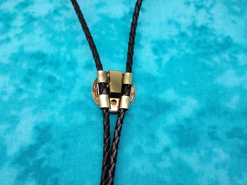 May include: A black braided bolo tie with a gold-toned metal clasp. The tie is on a turquoise background.