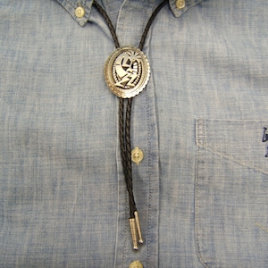 May include: A black bolo tie with a silver bolo tie slide featuring a stylized design. The bolo tie is worn on a light blue denim shirt.