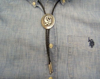 Könnte beinhalten: Ein schwarzes Bolo-Band mit einem silbernen Bolo-Band-Verschluss mit einem stilisierten Design. Das Bolo-Band wird auf einem hellblauen Jeanshemd getragen.