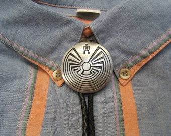 May include: A silver bolo tie with a circular pendant featuring a stylized maze design and a small figure in the center. The bolo tie is worn on a blue and orange striped shirt.