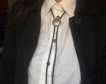May include: A person wearing a white button-down shirt with a black bolo tie. The bolo tie has a silver concho with a floral design.