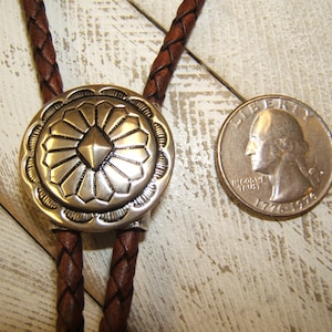 May include: A brown leather bolo tie with a silver, round, decorative tip featuring a geometric design. The tip is about the size of a US quarter.