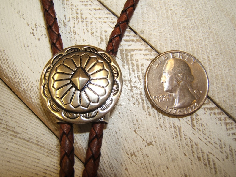 May include: A brown leather bolo tie with a silver, round, decorative tip featuring a geometric design. The tip is about the size of a US quarter.