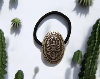 May include: Oval-shaped bronze-colored hair tie with a detailed embossed design. The hair tie features a black elastic band. The background includes green cacti and a white surface, creating a Southwestern aesthetic.