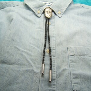 May include: A silver bolo tie with a decorative oval pendant and black braided cord. The bolo tie is displayed on a light blue button-down shirt. The shirt has a front pocket and a classic collar.