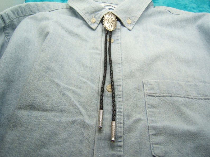 May include: A silver bolo tie with a decorative oval pendant and black braided cord. The bolo tie is displayed on a light blue button-down shirt. The shirt has a front pocket and a classic collar.