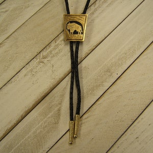 Puede incluir: Un bolo tie dorado con un diseño de búfalo tallado en el colgante. La corbata está hecha de cordón trenzado negro y tiene puntas doradas.