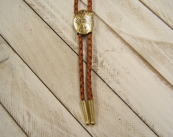 Könnte beinhalten: Ein braunes Leder-Bolo-Band mit einer goldfarbenen Metallspitze und einem dekorativen Blumenmuster.