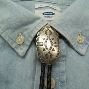 May include: A silver bolo tie with an oval, ornate pendant featuring geometric designs. The pendant is attached to a black braided leather cord. The tie is displayed on a light blue button-down shirt from Old Navy.