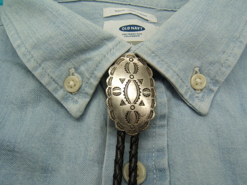 May include: A silver bolo tie with an oval, ornate pendant featuring geometric designs. The pendant is attached to a black braided leather cord. The tie is displayed on a light blue button-down shirt from Old Navy.