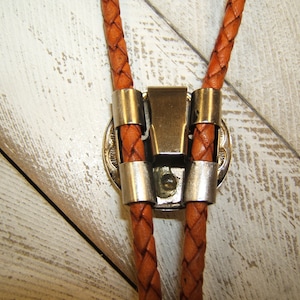 May include: A brown leather bolo tie with a silver clasp. The tie is braided and has a decorative metal piece on the clasp.