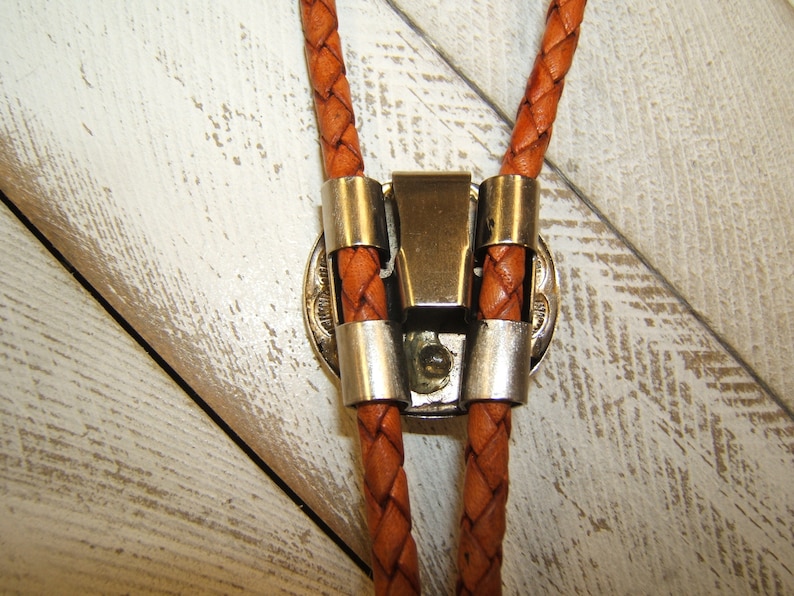 May include: A brown leather bolo tie with a silver clasp. The tie is braided and has a decorative metal piece on the clasp.