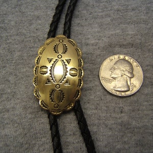 May include: A gold-colored bolo tie pendant with an oval shape and intricate etched design. The pendant is connected to a black braided cord. A quarter coin is next to the pendant for size comparison.