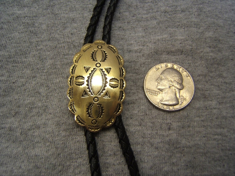 May include: A gold-colored bolo tie pendant with an oval shape and intricate etched design. The pendant is connected to a black braided cord. A quarter coin is next to the pendant for size comparison.