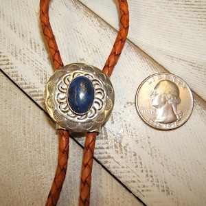 May include: A silver bolo tie with a blue stone set in a silver oval pendant. The bolo tie is on a brown leather cord.