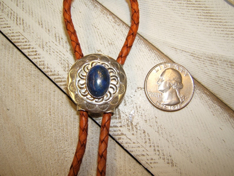 May include: A silver bolo tie with a blue stone set in a silver oval pendant. The bolo tie is on a brown leather cord.