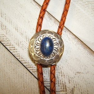May include: A bolo tie with a silver and blue stone pendant. The pendant has a circular design with a blue stone in the center. The bolo tie is made of brown leather.