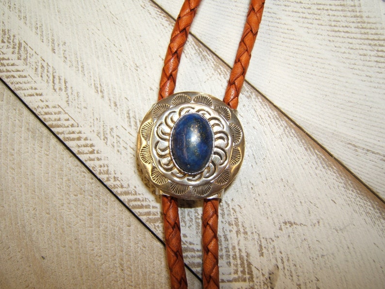 May include: A bolo tie with a silver and blue stone pendant. The pendant has a circular design with a blue stone in the center. The bolo tie is made of brown leather.