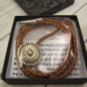 May include: A brown leather bolo tie with a silver and gold-toned metal tip featuring a geometric design. The bolo tie is in a black box with a white card that reads "the length of the P on Slide with your ms to Curl, you c e Cord. Also, ma helps to make in keepinrom se con"