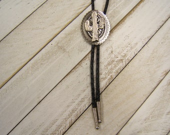 May include: A silver bolo tie with a cactus design. The tie is on a white wooden background.