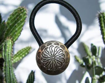 May include: A black elastic hair tie with a silver-toned metal button featuring a decorative floral design.