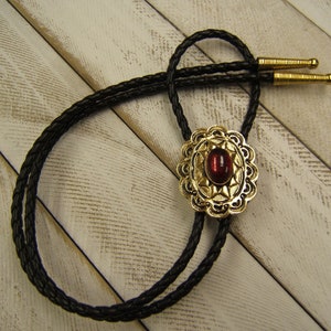 May include: A bolo tie with a braided black leather cord and gold-colored metal accents. The centerpiece features a red oval stone set in an ornate gold-colored filigree design. The tie has gold-colored tips.