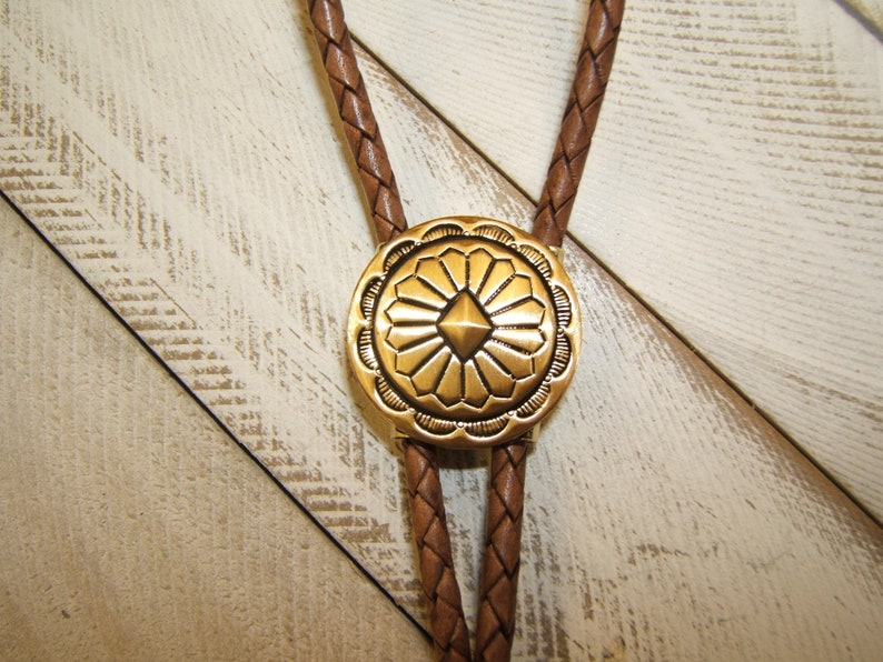 May include: A brown leather bolo tie with a gold-toned metal concho featuring a geometric design.