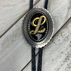 Pode incluir: Uma gravata bolo com um cordão trançado preto e um passador de metal prateado oval. O centro do passador contém um detalhe preto com uma letra L dourada adornada com strass transparentes, rodeada por uma borda decorativa recortada.