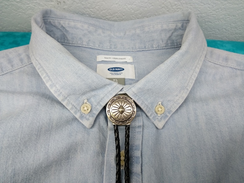 May include: A light blue denim shirt with a silver bolo tie featuring a floral design. The shirt has a button-down collar and a label that reads "Old Navy" and "XL".