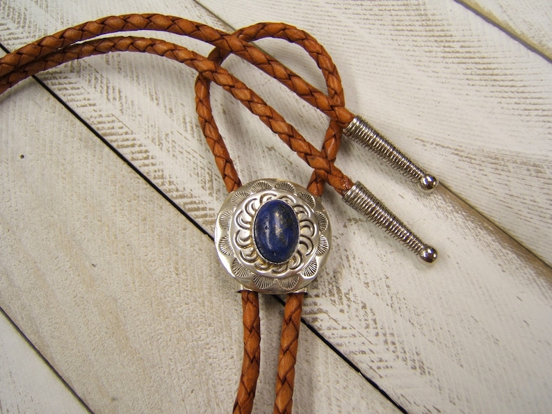 May include: A brown leather bolo tie with a silver and blue stone pendant. The pendant has a decorative design around the stone.