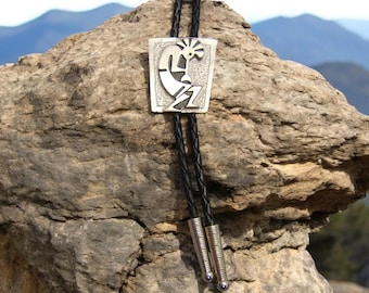 Op de afbeelding: Een zilveren bolo tie met een Native American Kachina-ontwerp. De bolo tie ligt op een rots met een berg op de achtergrond.