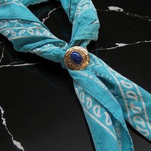 May include: A turquoise bandana with a white paisley pattern is secured with a gold-toned bolo tie clasp. The clasp features a dark blue oval stone in the center and is stamped with the words "MADE IN CHINA".