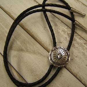May include: A black leather bolo tie with a silver, circular, decorative tip featuring a geometric design.