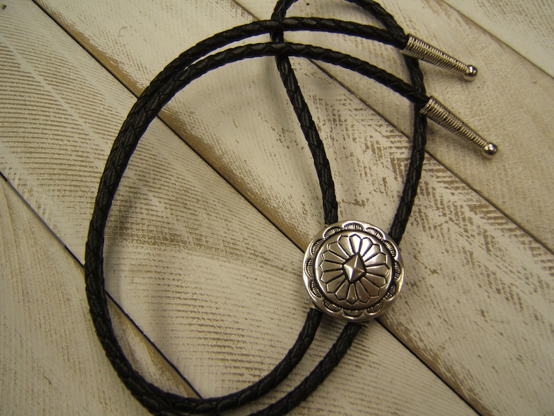 May include: A black leather bolo tie with a silver, circular, decorative tip featuring a geometric design.