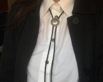 May include: A person wearing a white button-down shirt with a black bolo tie. The bolo tie has a silver concho with a floral design.